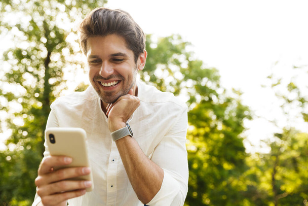Image of young caucasian brunette man smiling and holding cellphone while walking in park - Fotó, kép