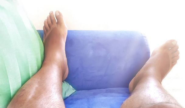 Τα πόδια ενός γυμνού άντρα που αναπαύεται σε έναν μπλε καναπέ με πράσινο μαξιλάρι. Κούραση και χαλάρωση στο σπίτι. Ξεκουράσου στον καναπέ. - Φωτογραφία, εικόνα