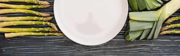 top view of fresh green leek, asparagus and empty plate on wooden surface, panoramic shot - Foto, Bild
