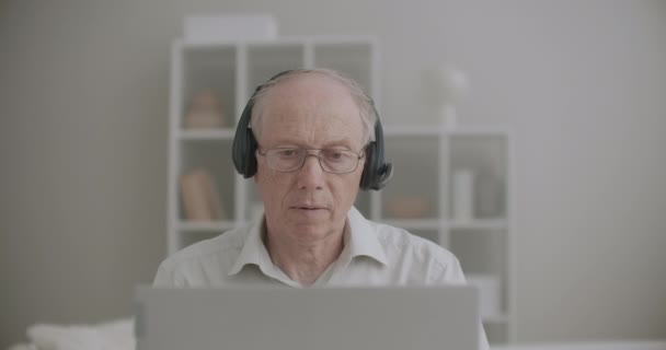 uomo anziano con le cuffie sta parlando e guardando la visualizzazione di notebook, conferenze online da casa - Filmati, video