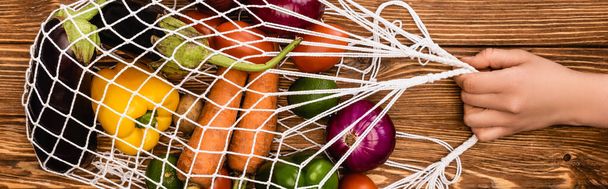 cropped view of woman holding string bag with fresh ripe vegetables on wooden table, panoramic shot - Photo, Image