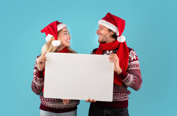 Feliz pareja joven en trajes de Navidad sosteniendo banner vacío con maqueta para el diseño sobre fondo azul - Foto, Imagen