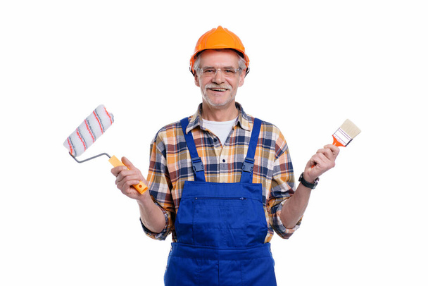 Photo of positive senior male builder holding paint roller and brush in hand, wearing special uniform, apron and protective helmet, isolated on white background. - Φωτογραφία, εικόνα