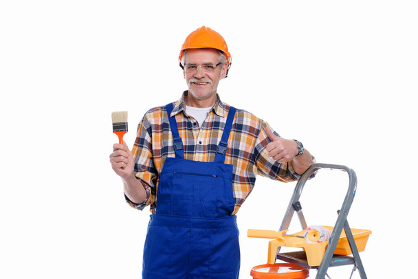 Professional builder, senior man in coverall and helmet ready for painting wall with roller, stay near stepladder. - Фото, зображення