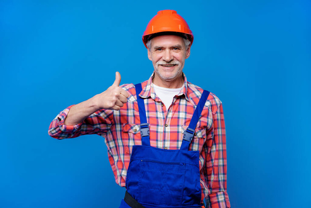 Smiling senior construction worker showing thumbs up isolated on blue background, repairment and decor concept. - Фото, зображення