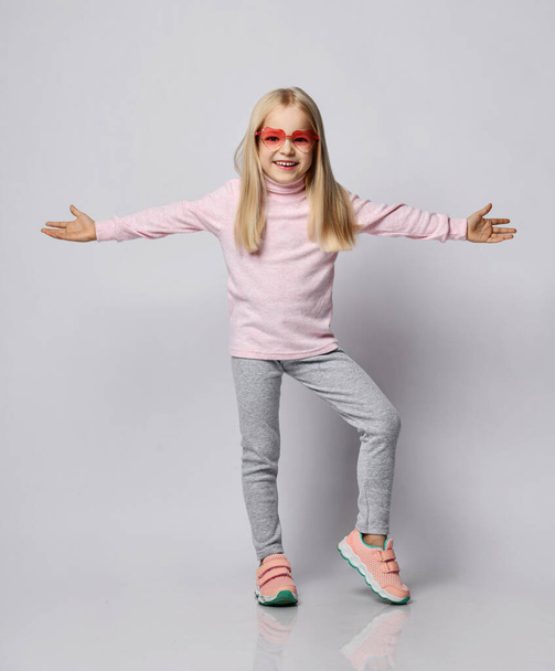 Blonde kid girl in stylish smoked heart-shaped sunglasses, turtleneck sweater and pants stands with arms spread wide - Fotó, kép