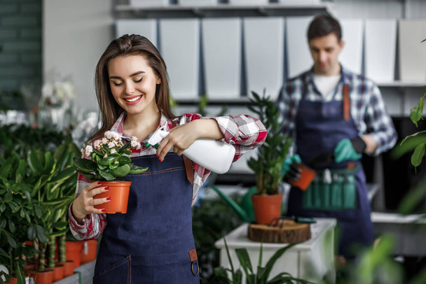 Gardeners concept, happy boy and girl with plants in beautiful garden center or Botanical center, pretty girl posing with beautiful flowerpot and sprayer. - Foto, afbeelding