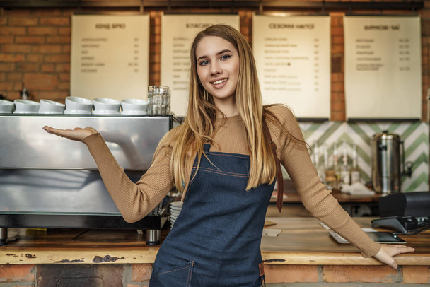 Nice stylish cheerful and attractive blonde girl, barista in casual shirt and apron, holding hand near bar counter. - Foto, immagini