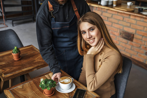 Barista man in blue apron giving order, latte with drawing for blonde woman customer in coffee shop, service mind concept. - Fotoğraf, Görsel