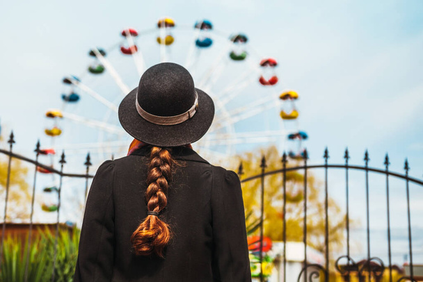 Вид ззаду на молоду дівчину в капелюсі, що стоїть перед коліщатком Феррі в парку розваг
. - Фото, зображення