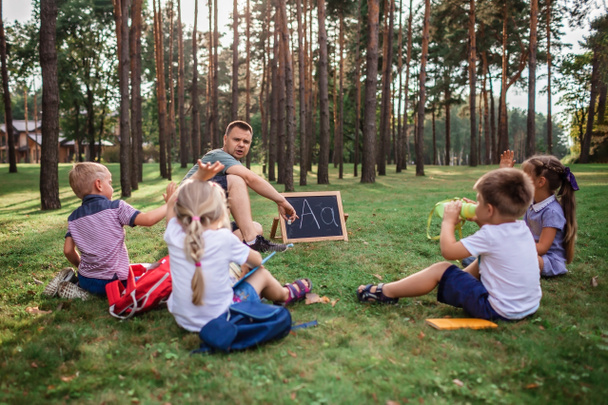 New normal back to school. Kindergarten and elementary scholars sitting with teacher on grass at open air class during pandemic. Safe hybrid education, social distance rule, new schooling guidance  - Valokuva, kuva