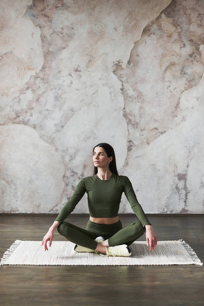 Young attractive woman with long dark hair, wearing sportswear, sitting in half lotus pose, resting after yoga practice, lokking away, indoor, full length, side view.Wellness concept.Advertising space - Photo, Image