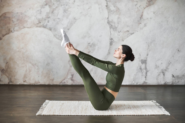 Sporty young woman with dark hair wearing sportswear doing stretching while sitting in Paripurna Navasana exercise, balance posture, working out in yoga studio against wall beige textured background - Fotó, kép
