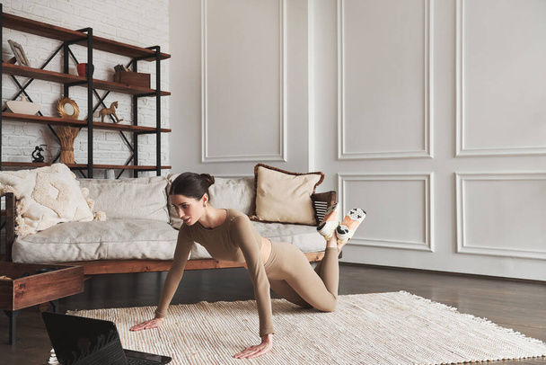 Young sporty woman working out at home interior, doing easy push ups exercise with knees on living room floor, using online fitness daily training program for beginners on laptop, side view. - 写真・画像