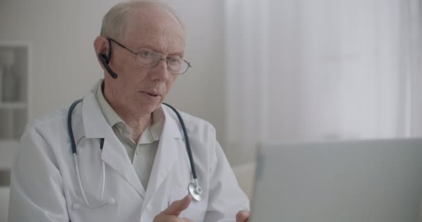 especialista médico de edad avanzada se está comunicando por cámara web en el ordenador portátil desde su oficina en la clínica, comunicación remota con los pacientes - Metraje, vídeo