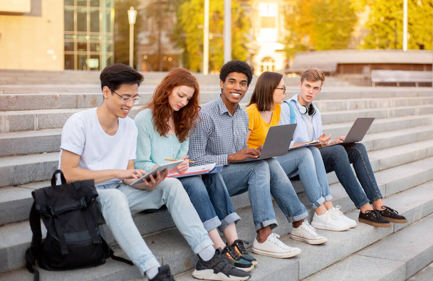 Többnemzetiségű diákok együtt tanulnak a kütyük használatával Ülve a szabadban - Fotó, kép