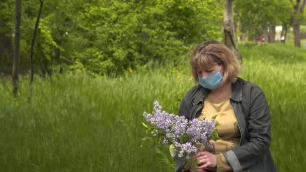 A nő a friss levegőn ül. Virágok vannak a kezében, orvosi maszk van az arcán. COVID-19 koronavírus világjárvány kitörésének koncepciója - Felvétel, videó