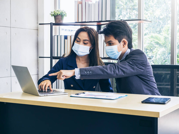 Ázsiai üzletember és nő öltönyt és védőmaszkot visel számítógép az asztalon, találkozó és együttműködés az irodában, új normális munkamódszer a koronavírus világjárvány lezárása után. - Fotó, kép