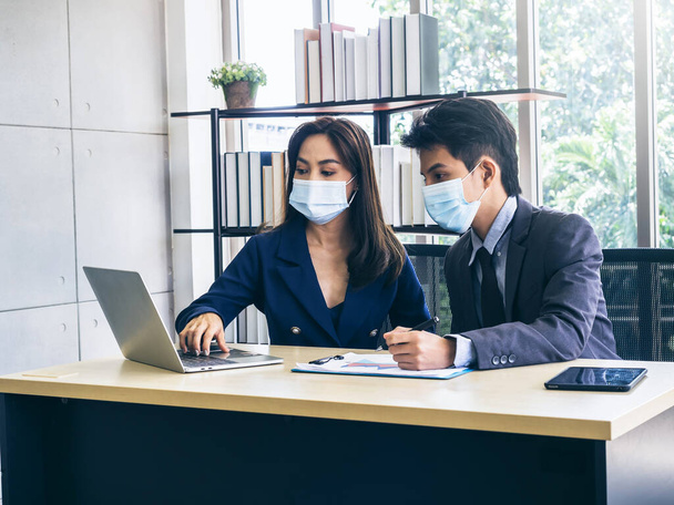Ázsiai üzletember és nő öltönyt és védőmaszkot visel számítógép az asztalon, találkozó és együttműködés az irodában, új normális munkamódszer a koronavírus világjárvány lezárása után. - Fotó, kép