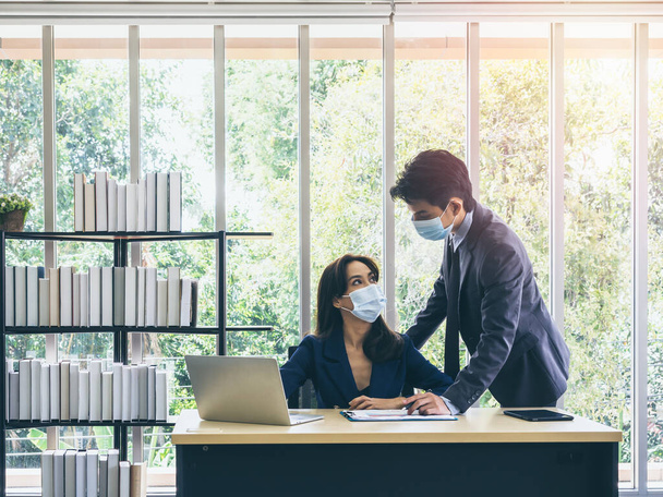 Homme d'affaires asiatique et femme portant costume et masques protecteurs en utilisant l'ordinateur sur le bureau, réunion et travailler ensemble au bureau, nouveau mode de vie normal après le verrouillage de la pandémie de coronavirus. - Photo, image
