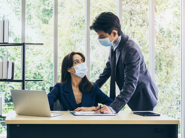 デスク上のコンピュータを使用してスーツや保護顔マスクを身に着けているアジアのビジネスの男性と女性,会議やオフィスで一緒に働く,コロナウイルスパンデミックのロックダウン後の新しい通常の作業ライフスタイル. - 写真・画像