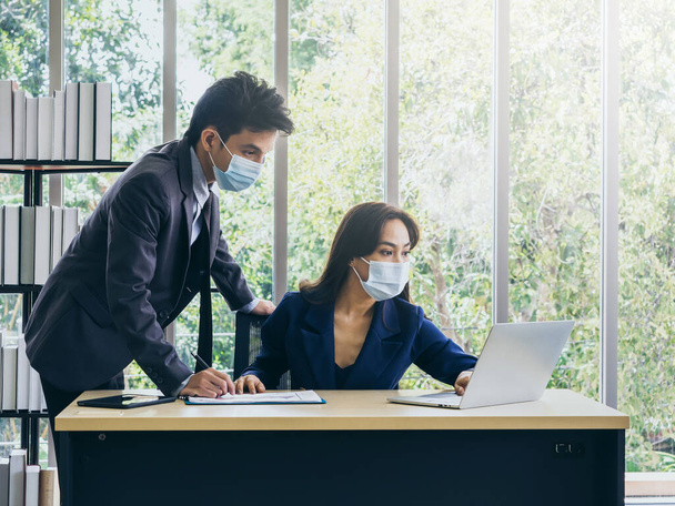 Uomo e donna d'affari asiatici che indossano tuta e maschere protettive usando il computer sulla scrivania, incontrandosi e lavorando insieme in ufficio, nuovo normale stile di vita di lavoro dopo il blocco pandemico coronavirus. - Foto, immagini