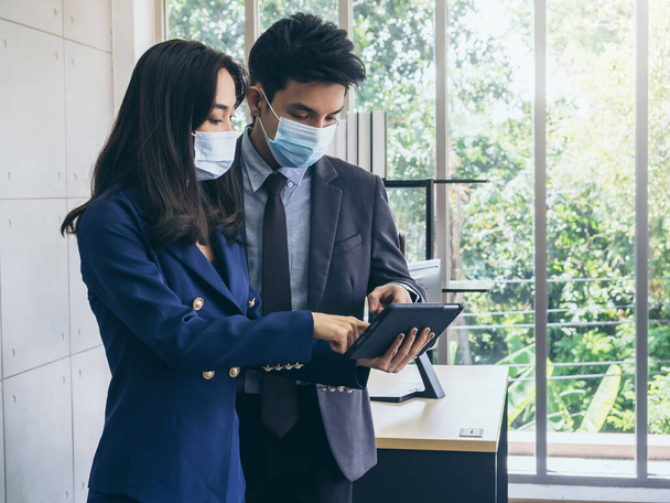 Asyalı iş adamı ve kadın takım elbise giyiyor ve koruyucu maskeler kullanıyor dijital tablet kullanıyor, ofiste buluşuyor ve birlikte çalışıyor, Coronavirus salgınından sonra yeni normal çalışma tarzı. - Fotoğraf, Görsel