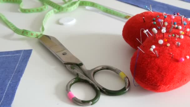 Naaiset, naaischaar, restjes blauwe stof, een rood speldenkussen met naalden in de vorm van een hartje, een centimeter op een witte tafelondergrond. Naaiatelier, workflow speelgoedfabriek - Video