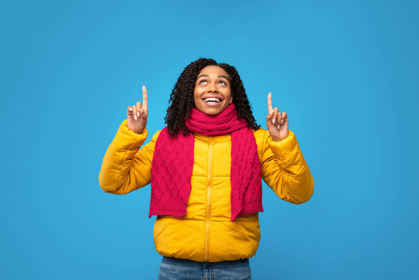 Black Woman In Jacket Pointing Fingers Upward Over Blue Background - Photo, Image