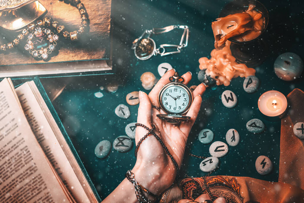 Astrology and esotericism. The female hand of the witch holds a watch on a chain. In the background, old books, fortune-telling runes, candles, and jewels. The view from the top. Light and dust. - Photo, Image