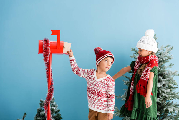 chlapec v klobouku a svetru dávat obálku do poštovní schránky v blízkosti sestry v zimním oblečení izolované na modré - Fotografie, Obrázek