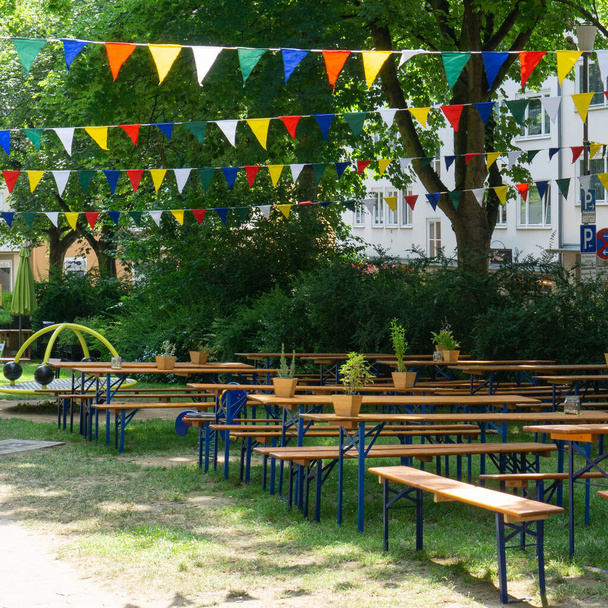 Άδειο τραπέζια και παγκάκια για ένα πάρτι στον κήπο το καλοκαίρι, χρωματιστές σημαίες απλώνονται ανάμεσα στα δέντρα - Φωτογραφία, εικόνα