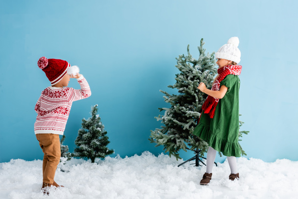 kids in winter outfit playing snowballs near christmas trees on blue - Photo, Image