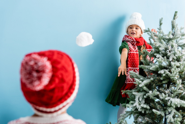 selective focus of girl in hat and scarf throwing snowball near brother on blue  - Photo, Image