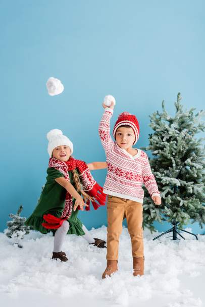 children in winter outfit playing snowballs near christmas trees on blue - Фото, изображение