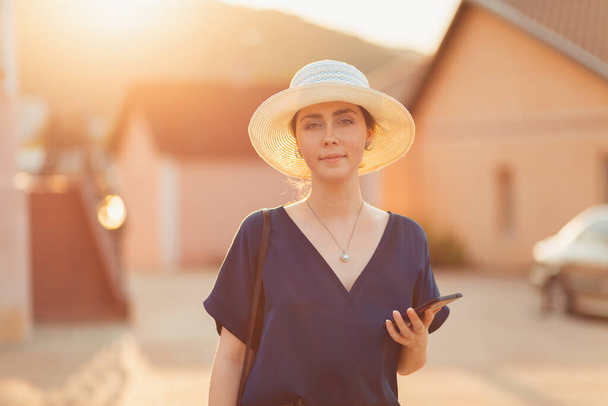 Comunicazione. Una donna elegante con un cappello sorride e tiene in mano uno smartphone. Sullo sfondo della casa e l'illuminazione al tramonto in una sfocatura. Il concetto di social networking, business e app per il telefono. - Foto, immagini