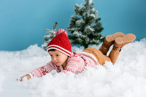 boy in winter outfit lying on white snow near christmas trees on blue - Foto, Imagen