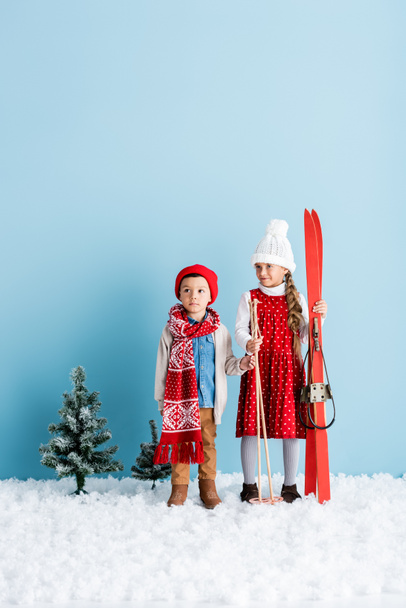 girl holding ski poles and skis while standing on snow near brother in winter outfit on blue - Foto, immagini