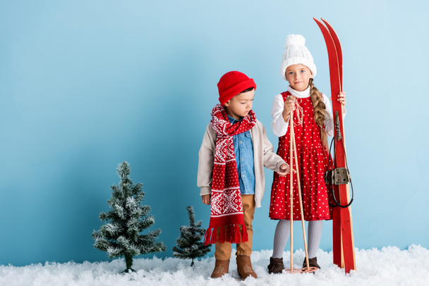 miúdo de chapéu e cachecol apontando com o dedo perto da irmã segurando bastões de esqui e esquis, enquanto em pé no azul - Foto, Imagem