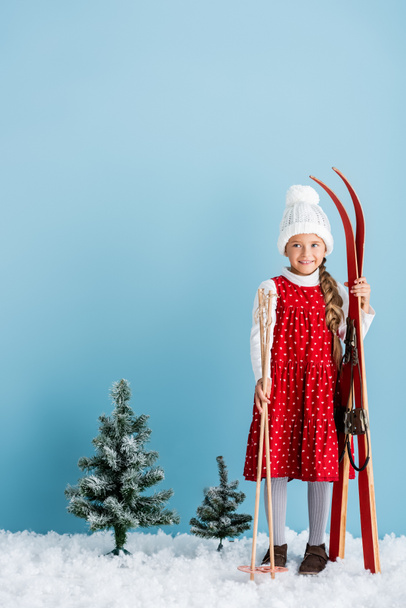 kid in winter outfit standing with ski poles and skis on blue - 写真・画像