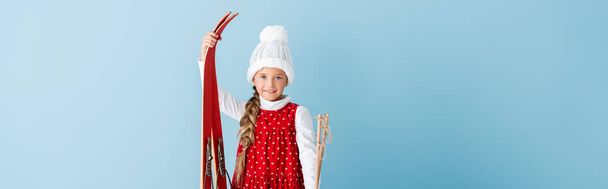 horizontal image of kid in winter outfit standing with ski poles and skis isolated on blue - Photo, Image