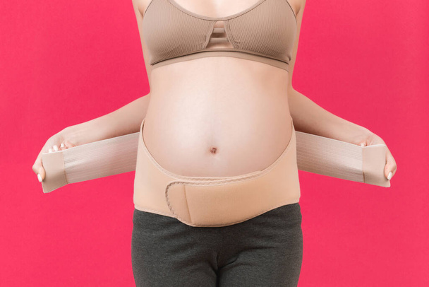 Primer plano de la mujer embarazada vistiendo banda de maternidad elástica en el fondo rosa con espacio de copia. Concepto de cinturón de soporte abdominal ortopédico. - Foto, Imagen