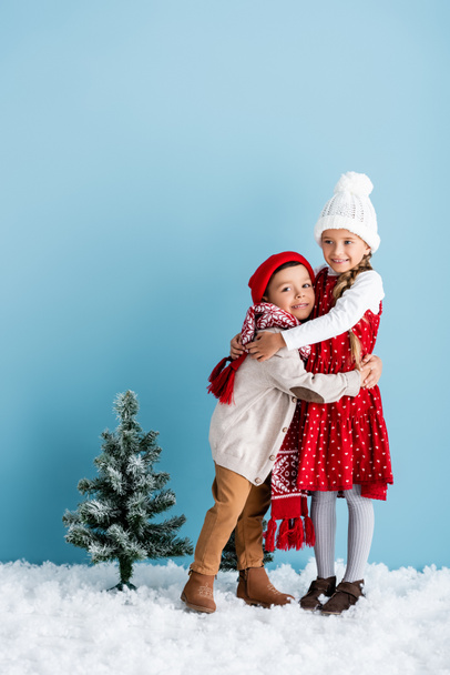 Kinder im Winteroutfit umarmen sich neben dem Weihnachtsbaum auf blauem Grund - Foto, Bild