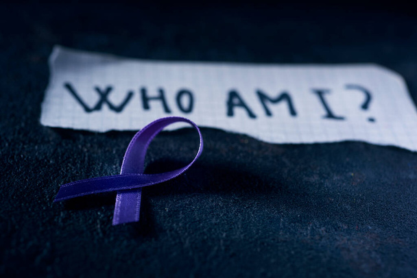 una cinta de conciencia púrpura, para la conciencia de la enfermedad de Alzheimers, y un pedazo de papel con la pregunta ¿quién soy yo escrito en ella, en una superficie de textura negro - Foto, Imagen