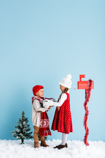 Сестра и брат в зимнем наряде смотрят друг на друга, держа в руках подарок возле почтового ящика на голубом - Фото, изображение