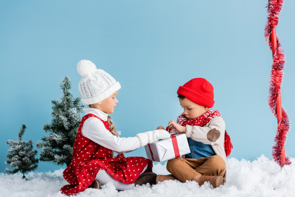garçon et fille en tenue d'hiver assis sur la neige et touchant présent isolé sur bleu - Photo, image