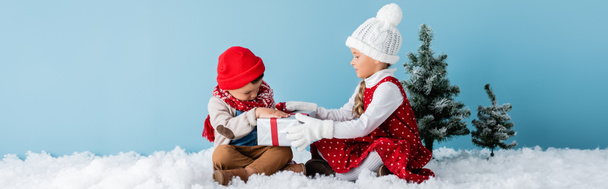 Panoramaaufnahme von Schwester und Bruder im Winteroutfit auf Schnee sitzend und anrührendes Geschenk auf Blau - Foto, Bild