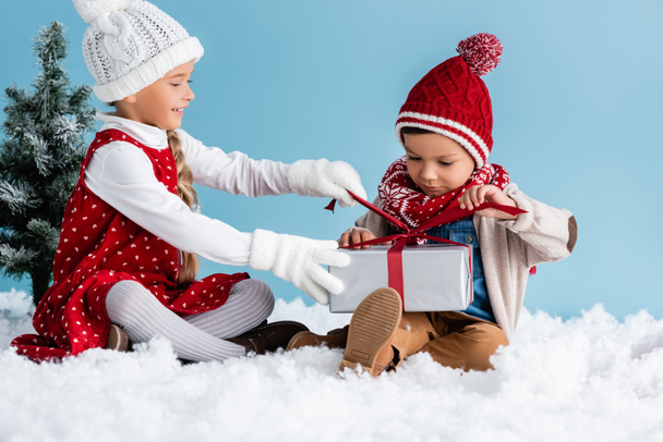 Kinder in Hüten und Winterbekleidung sitzen auf Schnee und berühren Geschenk isoliert auf blau - Foto, Bild