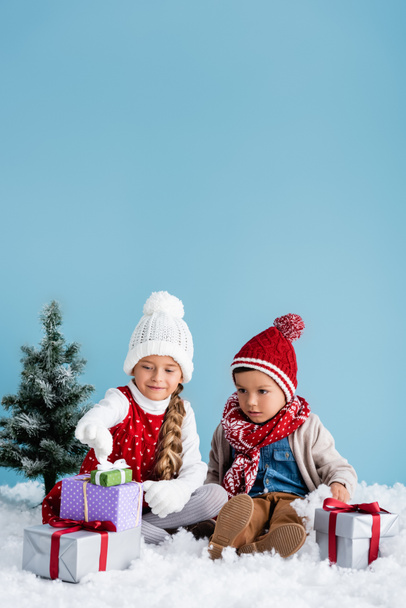 Kinder im Winteroutfit sitzen auf Schnee und betrachten Geschenke in der Nähe von Tannenbäumen isoliert auf blauem Grund - Foto, Bild