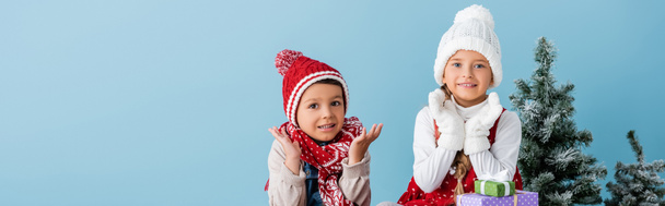 plano panorámico de los niños en traje de invierno gesto cerca de regalos y abeto aislado en azul - Foto, imagen
